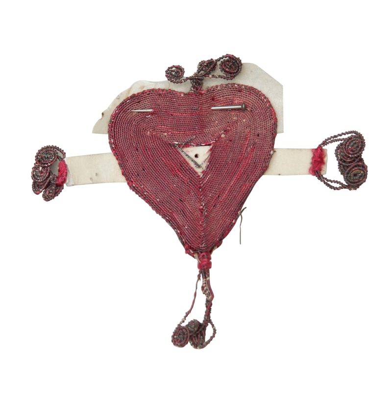 A red embroidered heart with a pin stuck horizontally through the top. It has embroidered scrolls at the top and bottom, and at the ends of paper 'arms' extending from its sides.